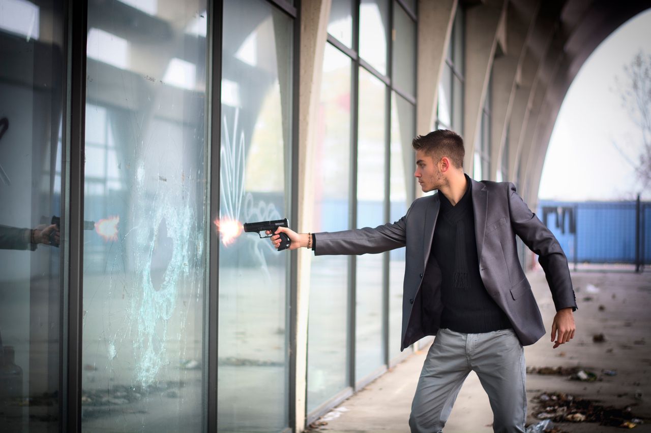 A staged scene: a man in a suit fires a handgun at a wall of storefront glass, which has cracked around the impact point but is still standing.