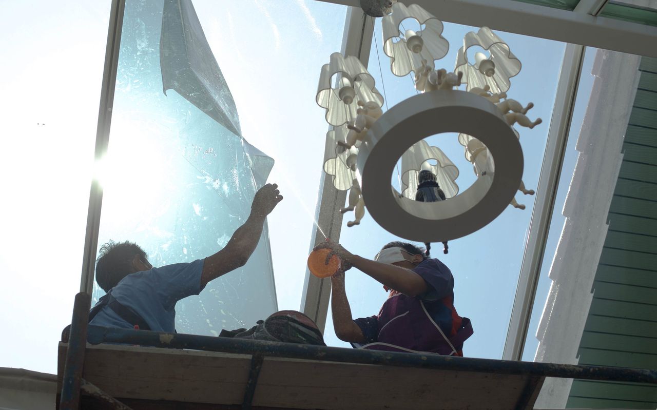 Two installers on a work platform applying film to a large, high glass panel, one smoothing the film with a squeegee.