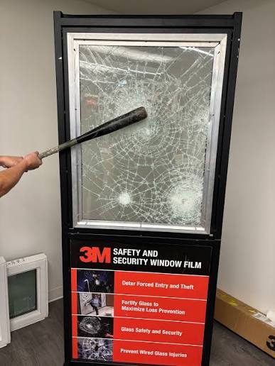 A 3M safety and security window film demonstration stand. A framed pane is being struck with a baseball bat and has crazed into a dense web of cracks without leaving the frame.
