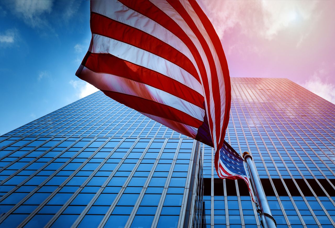 An American flag flying in front of a tall glass office tower, seen from below against a blue sky.