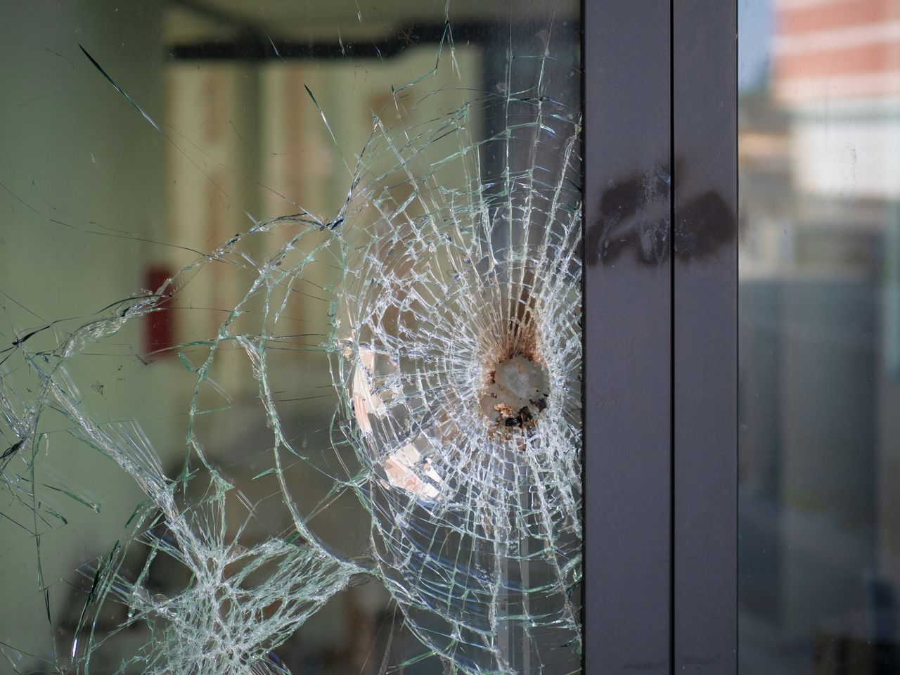 Close-up of a glass pane beside a dark metal frame with a single round lodged in it. Cracks radiate out in a wide web, and the pane is still in place.
