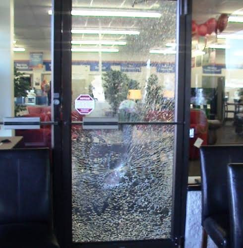 A retail storefront door photographed from inside. The full glass panel is crazed into small fragments across its whole area but remains standing in the door frame.
