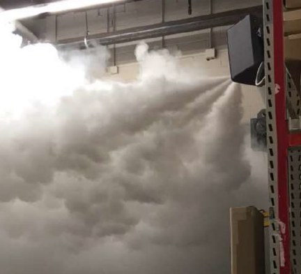 A wall-mounted fog head discharging along a warehouse ceiling, the barrier already spreading across the full width of the aisle.